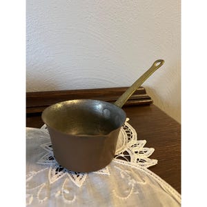 May include: A small, vintage copper saucepan with a brass handle. The interior of the pan is a darker color than the exterior. The handle has a hole at the end for hanging. The saucepan is sitting on a white lace doily.