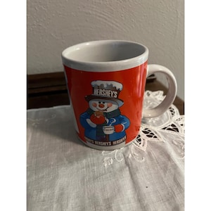 May include: A red Hershey's cocoa mug featuring a snowman wearing a hat and scarf, holding a mug of cocoa. The mug has a white handle and the Hershey's logo is visible.