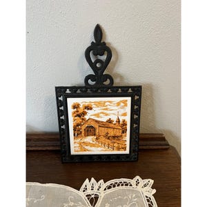 May include: Black cast iron trivet with a square tile featuring a sepia-toned image of a covered bridge. The trivet has a decorative handle and a patterned border. A vintage home decor item.