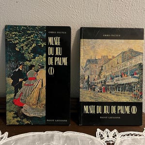 May include: Two vintage books titled "Musée Du Jeu De Paume" with the text "Orbis Pictus" and "Payot Lausanne" on the covers. The book covers feature illustrations of people and buildings. The books are black.