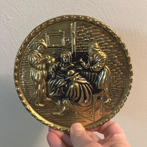 May include: A round, gold-colored metal relief plate depicting a scene with several figures in period clothing. The plate has a detailed, raised design and a decorative, textured border. The figures are arranged around a central seated figure.