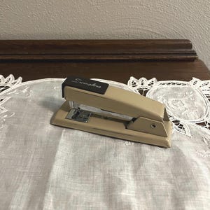 May include: A tan and brown Swingline stapler. The stapler is on a white lace doily. The word "Swingline" is printed on the top of the stapler in black. The stapler is made of metal and plastic.