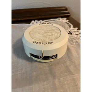 May include: A vintage white Westclox alarm clock with a round face and a rotating dial. The clock face has the words "LONG RING" and the brand name "WESTCLOX". The dial shows the number "55". The clock is set on a white lace doily.