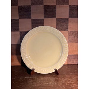 May include: A pale yellow ceramic plate with a decorative leaf pattern around the rim. The plate is round and sits on a small wooden stand, set against a checkered brown and tan background. A simple, elegant design for serving food.