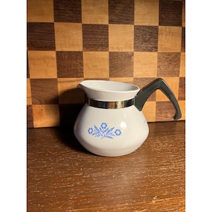 May include: A white ceramic creamer pitcher featuring a blue floral pattern on the front. The pitcher has a metallic silver band around the neck and a black angular handle extending from the side. The design includes a wide spout for pouring.