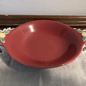 May include: A vintage, red-toned ceramic bowl with a shallow design and two handles. Each handle features a yellow floral design with green leaves. The bowl is likely for serving or display.