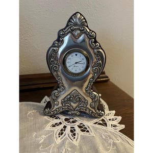 May include: A silver-toned decorative mantel clock with an ornate, scrolling design. The clock face is white with black numerals and hands. The clock is set on a dark brown base and sits on a white lace doily.