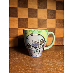 K&ouml;nnte beinhalten: Ein hellgr&uuml;ner Keramikbecher mit einer stilisierten Illustration der Figur Stitch mit gro&szlig;en dunklen Augen und einer blauen Nase. Der Becher hat einen glatten Henkel und ein hellgr&uuml;nes Inneres, mit einem Muster aus gr&uuml;nen Formen um die Figur herum.