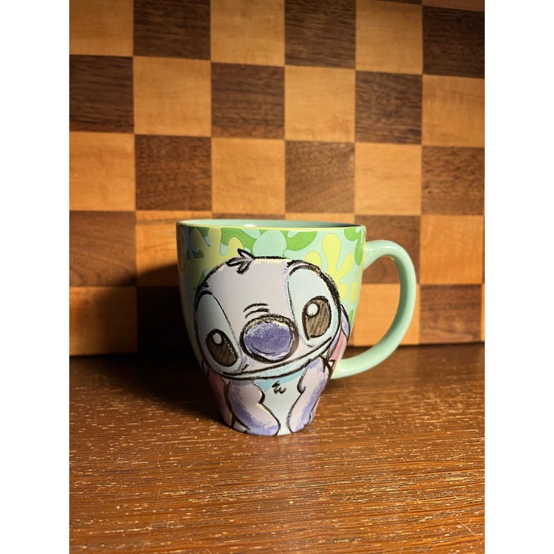 K&ouml;nnte beinhalten: Ein hellgr&uuml;ner Keramikbecher mit einer stilisierten Illustration der Figur Stitch mit gro&szlig;en dunklen Augen und einer blauen Nase. Der Becher hat einen glatten Henkel und ein hellgr&uuml;nes Inneres, mit einem Muster aus gr&uuml;nen Formen um die Figur herum.