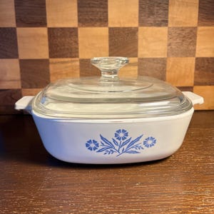 May include: A white casserole dish with a clear glass lid. The dish features a blue floral design on the front. The lid has a clear glass knob. The dish has two white handles on either side.