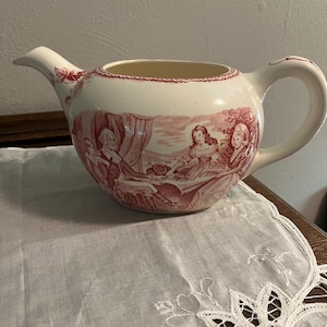 Puede incluir: Cremera de cerámica color crema con un diseño rojo. La cremera tiene un pico, un asa y una escena decorativa de figuras en un paisaje. El borde está adornado con un borde rojo. La cremera está sobre un paño blanco bordado.