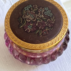Op de afbeelding: Een vintage, ronde, paarse glazen pot met een decoratief deksel. Het deksel is bruin met een geborduurd bloemenontwerp en een goudkleurige metalen rand. De pot heeft een geribbelde textuur.