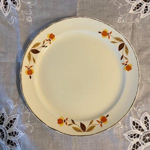 May include: A white ceramic dinner plate with a gold rim and a brown, orange, and yellow floral design.