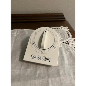 May include: A white Cooks Club kitchen timer with a dial and a large, white, triangular timer. The timer has a black dial with numbers and hash marks. The text "Cooks Club QUALITY & DESIGN" is printed on the front.