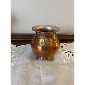 May include: A small, copper-colored metal vase with a silver-colored rim and a decorative emblem. The vase has a rounded body and sits on a gold-colored, ornate base. The emblem features a detailed design.