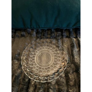May include: A clear glass plate featuring a textured hobnail pattern across the surface and a central sunburst design. The plate has a scalloped edge and a transparent finish, showcasing a circular arrangement of raised glass dots.