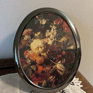 May include: Oval-shaped picture frame with a dark metallic finish. The frame holds a floral still life painting in shades of red, orange, yellow, and white. The frame has small ball feet for support, and is displayed on a white lace cloth.
