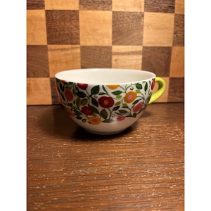 May include: A white ceramic teacup with a floral pattern of red, orange, and pink flowers with green leaves. The handle is bright yellow. The cup is sitting on a wooden surface.