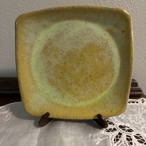 May include: Square ceramic plate with rounded corners. The plate has a speckled, glazed surface with a gradient of colors, transitioning from a light green center to a golden-yellow rim. The plate is displayed on a wooden stand.