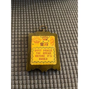 May include: A rectangular wooden wall plaque with a dark green frame and a yellow center featuring cross-stitched images of wheat, bread, and a basket. The text reads: DONT PRAISE THE BREAD BEFORE ITS BAKED. It includes a brass hanging loop.