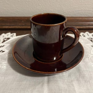 May include: A brown ceramic coffee cup and saucer set. The cup has a rounded handle and the saucer has a slightly raised edge.