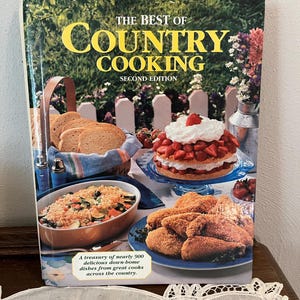 May include: A cookbook titled "The Best of Country Cooking" with a cover featuring a garden scene and various dishes. The book includes images of bread, a strawberry shortcake, fried chicken, and a casserole. The text reads "Second Edition."