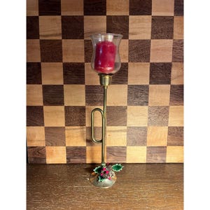 May include: A brass trumpet-shaped candle holder featuring a tall, slender stem and a clear glass cup at the top containing a red wax candle. The base is decorated with green holly leaves, small red berries, and two small brown pinecones.