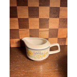 May include: A cream-colored ceramic creamer featuring a speckled finish and a handle on the right side. The exterior is decorated with a horizontal blue band above a floral pattern consisting of yellow flowers and small blue and green botanical sprigs.