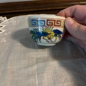 May include: Small, white ceramic bowl with a colorful bird design. The bowl features a repeating geometric pattern in red, blue, and brown around the rim. The bird design includes yellow, blue, green, and red details.