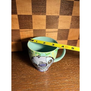 K&ouml;nnte beinhalten: Ein hellblauer Keramikbecher mit einer Grafik der Figur Stitch auf der Vorderseite. Ein gelbes Ma&szlig;band liegt &uuml;ber dem oberen Rand und zeigt einen Durchmesser von etwa 8,9 cm f&uuml;r die &Ouml;ffnung des Bechers an.