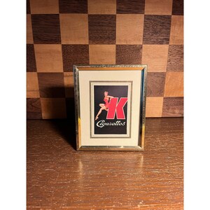 May include: A rectangular gold-toned metal frame containing a vintage-style advertisement print. The print features a large red letter K, a small illustration of a person in a red outfit, and the white text Cigarettes on a black background.