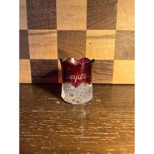 May include: A small, ruby red glass cup with a scalloped rim and a clear, diamond-cut base. The word "Comforia" is written in cursive on the red glass. The cup is likely vintage and decorative.