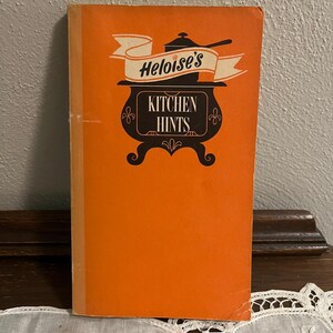 May include: An orange cookbook titled "Heloise's Kitchen Hints" with a black and white illustration of a pot and banner. The book is standing upright on a white lace tablecloth.