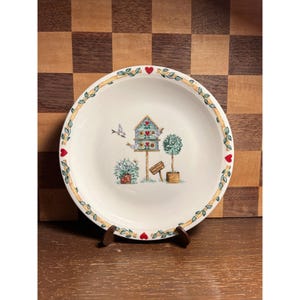 May include: A white ceramic plate with a decorative birdhouse scene. The plate features a birdhouse, two topiary trees, and a small flower pot. The rim is adorned with red hearts and green vines. The plate is displayed on a wooden stand.