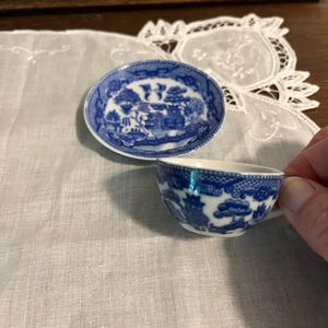 May include: A small blue and white teacup and saucer set. The set features a classic Willow pattern design. The cup is held by a hand. The saucer is oval-shaped. The set is placed on a white cloth.