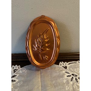 May include: A copper-colored, oval-shaped mold with a leaf design. The mold has a decorative, wavy edge and a small loop at the top for hanging. The leaf is detailed with veins and a small acorn at the bottom.