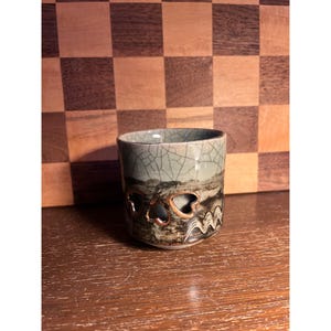 May include: A cylindrical ceramic cup featuring a light green crackle glaze on the upper portion and a dark brown base with cutout heart shapes. The lower section displays a landscape-inspired pattern with wavy lines and metallic-toned accents.