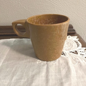 May include: A tan ceramic mug with a rounded body and a square handle. The mug has a speckled texture and sits on a white lace doily. The mug is empty and ready to be filled with a warm beverage.