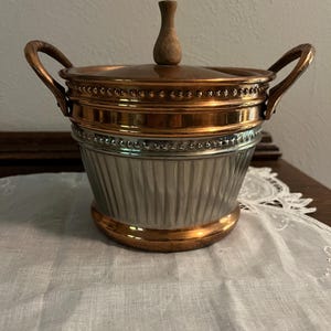 May include: A vintage copper and silver-toned ice bucket with a wooden handle on the lid and two copper handles. The bucket has a fluted silver-toned body with copper accents and a copper rim at the base.
