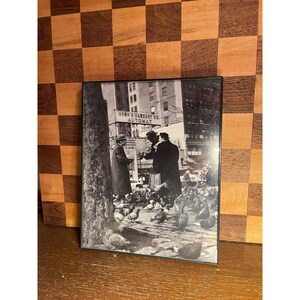 May include: A black and white framed photograph depicting a city street scene with two people standing near a tree and a large flock of pigeons on the ground. A sign above reads HORN & HARDART CO. AUTOMAT, with tall buildings visible in the background.