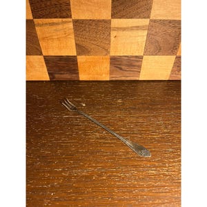 May include: A slender silver-toned cocktail fork featuring three thin tines and a decorative floral motif embossed on the handle. The metal utensil has a polished finish and a long, narrow stem, designed for serving small appetizers or garnishes.