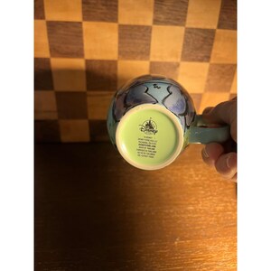 K&ouml;nnte beinhalten: Ein hellblauer Keramikbecher mit einem Disney-Logo auf der Unterseite. Der Boden ist hellgr&uuml;n und zeigt den Text: Disney Store USA, LLC, Pasadena, CA 91105, DISNEYSTORE.COM, MADE IN THAILAND, FABRIQUE EN THAILANDE, HECHO EN TAILANDIA.