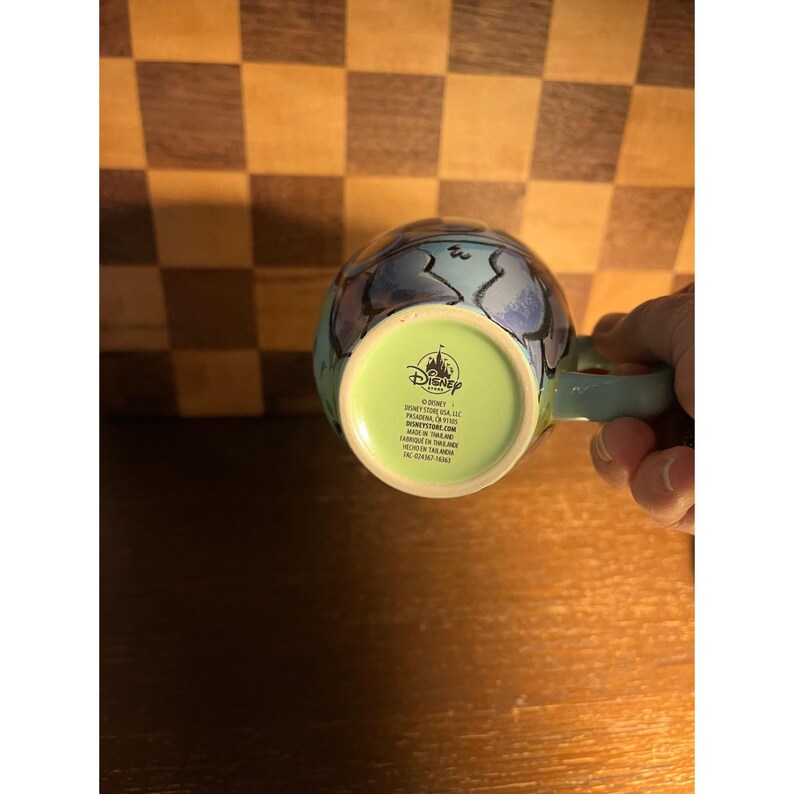 K&ouml;nnte beinhalten: Ein hellblauer Keramikbecher mit einem Disney-Logo auf der Unterseite. Der Boden ist hellgr&uuml;n und zeigt den Text: Disney Store USA, LLC, Pasadena, CA 91105, DISNEYSTORE.COM, MADE IN THAILAND, FABRIQUE EN THAILANDE, HECHO EN TAILANDIA.