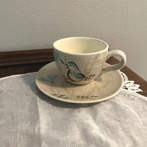 Könnte beinhalten: Ein Keramik-Tee-Tassen- und Untertassen-Set. Die Tasse und die Untertasse sind beige mit einem gesprenkelten Muster. Auf der Tasse und der Untertasse ist ein blauer Vogel aufgemalt. Die Tasse hat einen gebogenen Henkel.