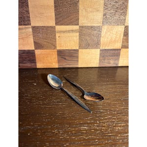 May include: Two vintage silver spoons with ornate handle detailing. The spoons are placed on a dark wooden surface, with a checkered wooden background. The spoons have a classic, elegant design.