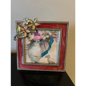 May include: A red picture frame with a floral design. The frame has a silver border and a colorful floral embellishment on the top left corner. The picture inside the frame depicts a vase of flowers and a duck.