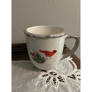 May include: White ceramic mug with a gray rim and handle, featuring a red cardinal bird perched on a branch with berries. The mug has a rustic, textured handle and rim, and is set on a white lace doily.