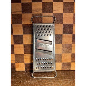 May include: A rectangular metal kitchen grater featuring a top and bottom handle, a series of small circular perforations, and two horizontal slicing blades. The center of the grater is embossed with the text BROMCO MADE IN U.S.A.