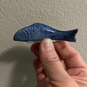 May include: A blue ceramic fish-shaped chopstick rest. The fish has detailed scales and fins, and is held in a hand. The chopstick rest is a decorative item for the dining table.