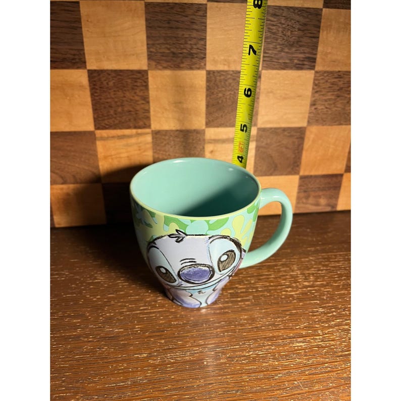 K&ouml;nnte beinhalten: Ein hellblauer Keramikbecher mit einer Zeichentrickfiguren-Illustration auf der Vorderseite. Ein gelbes Ma&szlig;band ist hinter dem Becher positioniert und zeigt eine H&ouml;he von etwa 10 cm vom Boden bis zum oberen Rand des Bechers an.